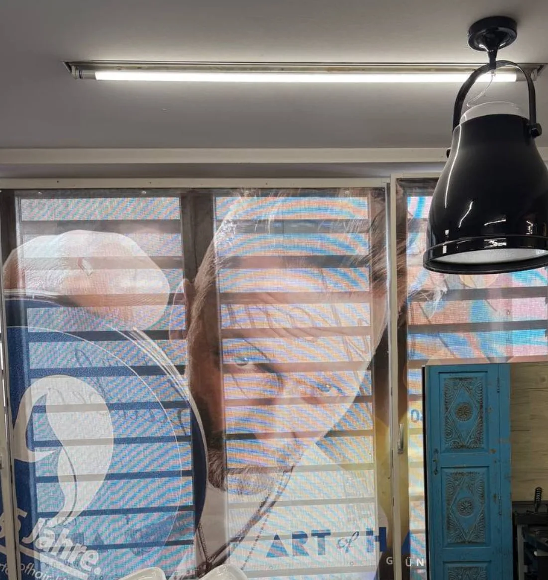 Fensterfront der Lounge, bespannt mit der Jubiläumsfolie, darüber eine schwarze Industrieleuchte, rechts eine türkise Holztür