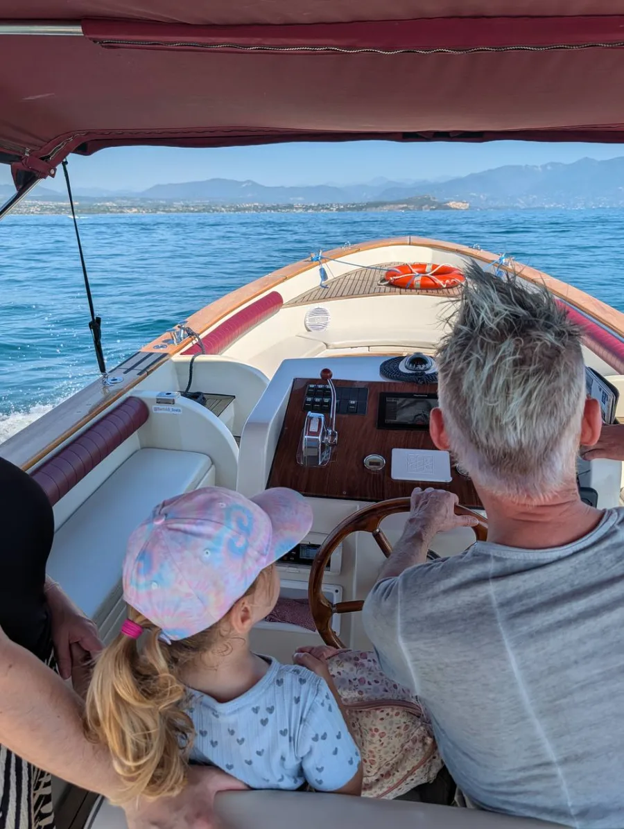 Günther Fischer am Steuer eines Holzboots auf einem See, von hinten; neben ihm ein Kind mit bunter Kappe